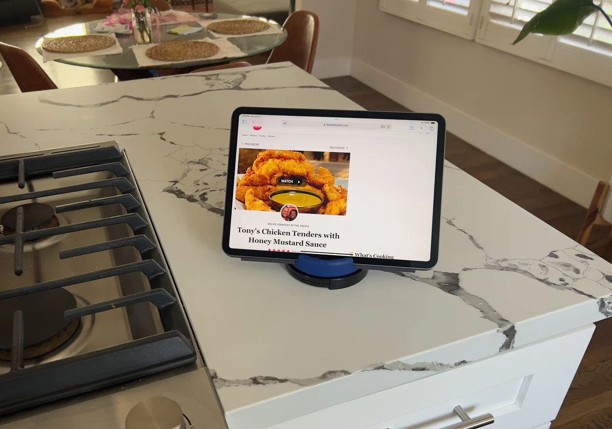 A tablet propped at reading height by a blue stand on a marble kitchen counter beside a stove